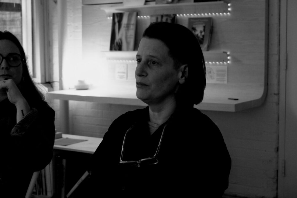 Karen Vanderborght assise lors d’un atelier, photographiée en noir et blanc, avec des lunettes suspendues au cou et des étagères en arrière-plan