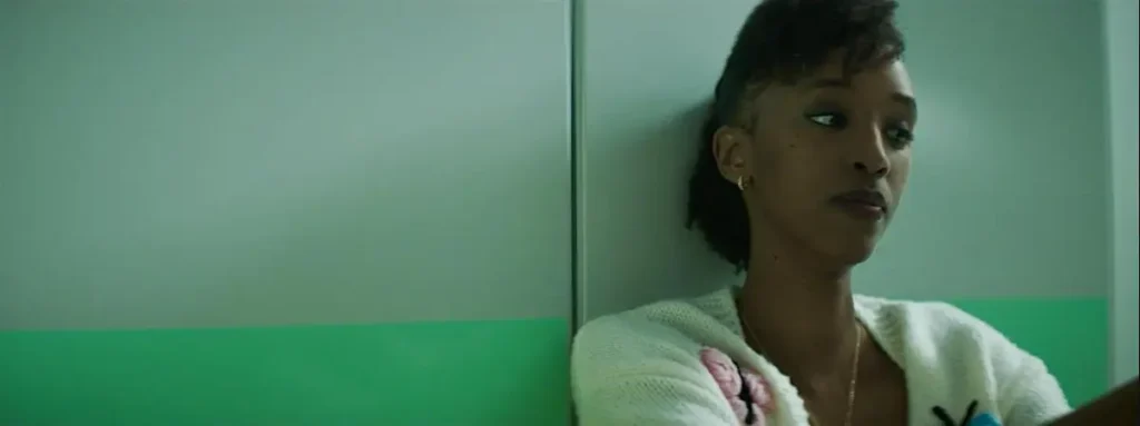 A young woman leaning against a green and white wall looks into the distance, in an introspective scene from the film Rêver en néon.
