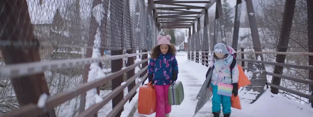 Two children walk across a snowy bridge carrying colorful suitcases, in a scene from the film Rêver en néon.