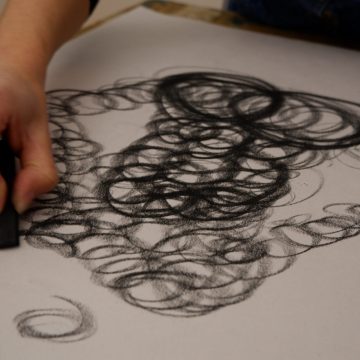 Main dessinant des cercles au fusain sur une feuille blanche.